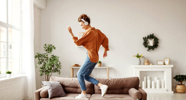 Cheerful woman listening to music and dancing on soft couch at home in day off Positive young lady in wireless headphones and casual clothes listening to music with pleasure and dancing of sofa in light modern living room lifestyle people stock pictures, royalty-free photos & images