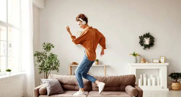 Positive young lady in wireless headphones and casual clothes listening to music with pleasure and dancing of sofa in light modern living room