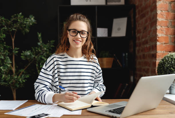 istock Happy millennial woman taking notes in notepad while working at laptop in comfortable loft office 1219638472