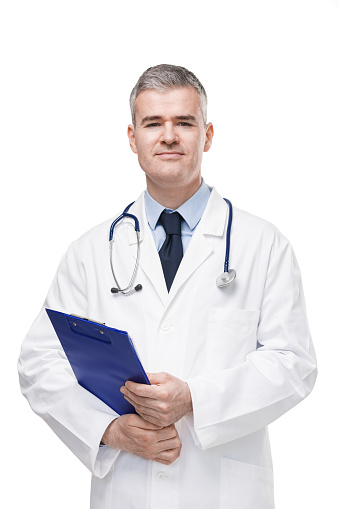 Doctor in lab coat and stethoscope holding a patient file or medical notes on a clipboard in his hands as he turns to look at the camera isolated on white