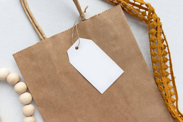 istock Blank white gift luggage tag on a brown paper bag - Flat lay gift tag mockup 1219661785