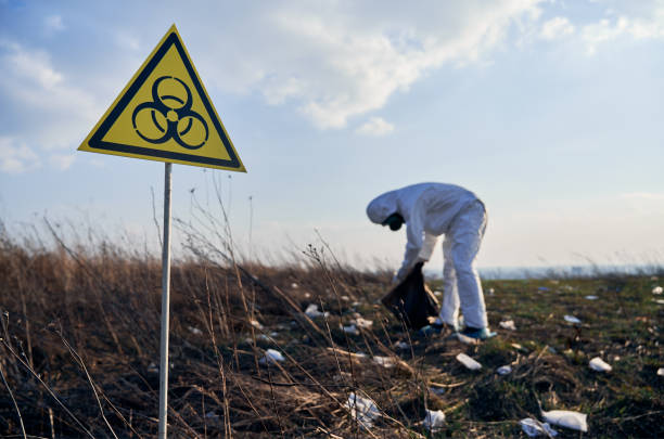 Focus on biohazard sign. On the blurred background researcher wearing white protective coverall, gas mask, collecting plastic garbage into black waste bag outdoors on a sunny day in spring