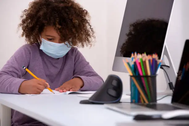 Six years old schoolgirl studying remotely far away from her classmates during a global epidemic. The girl sitting at a table, writing with a pencil on an exercise book, using a computer, and the internet. The child wearing a surgical mask, keeping herself protected from the virus. Coronavirus pandemic education concept.