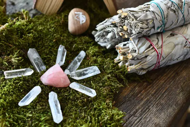 A close up image of a rose quartz healing crystal grid on green moss with white sage smudge sticks.