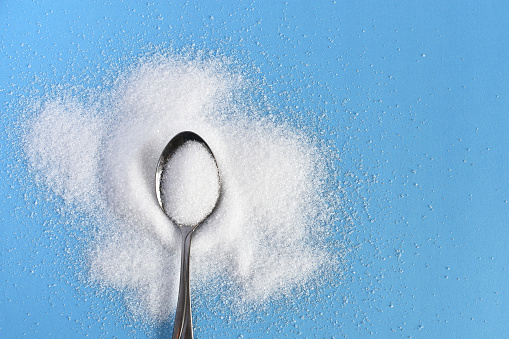 A top view image of a metal spoon and white sugar on a bright blue background.