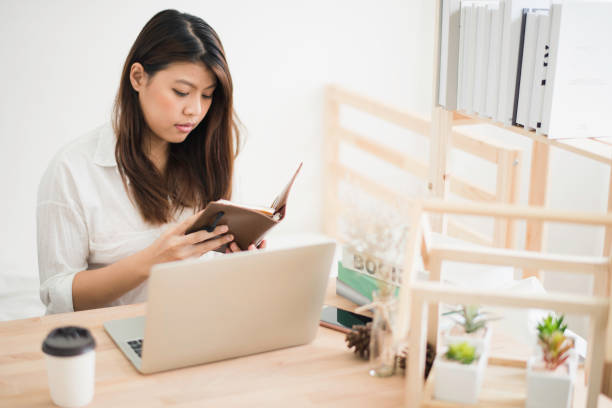 istock Young beautiful Asian woman sitting on white bed working with computer laptop which put on wood table and read the note book. Woman glad and smiling with felling happy. Work at home office concept 1219685634