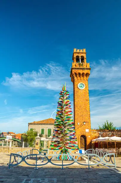 Murano clock tower Torre dell'Orologio of San Stefano church, Colorful christmas tree made of Murano Glass on Campo Santo Stefano square, Murano islands, Veneto Region, Northern Italy, Vertical view