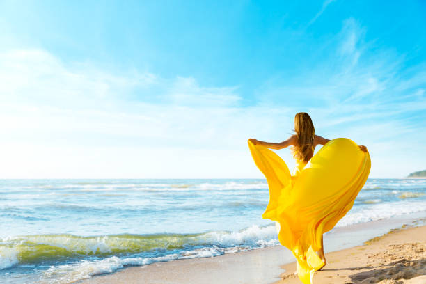 woman on sunny sea beach in yellow fluttering dress, fashion model back rear view, silk cloth waving on wind - activity baltic countries beauty in nature blue imagens e fotografias de stock