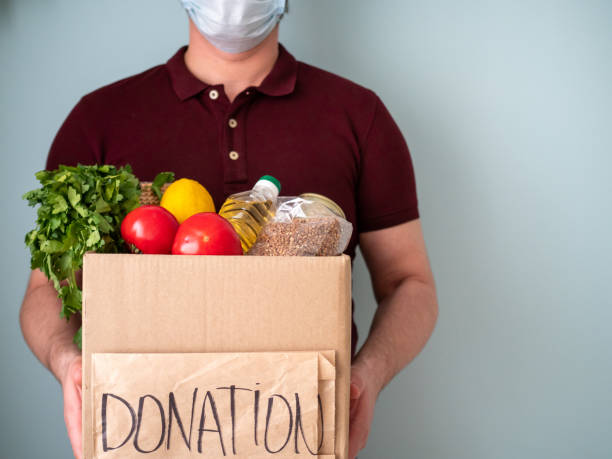 Man keeps donation box with goods in his hands, neighbourhood help concept Caucasian man in face mask keeps donation box with goods and food in his hands on blue background, neighbourhood help concept at quarantine time because of coronavirus infection, copy space transparent donation box stock pictures, royalty-free photos & images