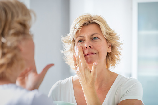 Mujer adulta media extendiendo una capa de crema facial frente al espejo photo