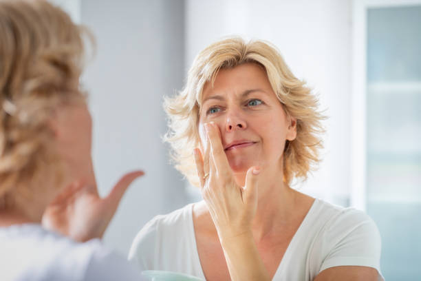 mujer adulta media extendiendo una capa de crema facial frente al espejo - skin care blue eyes expressing positivity cheerful fotografías e imágenes de stock