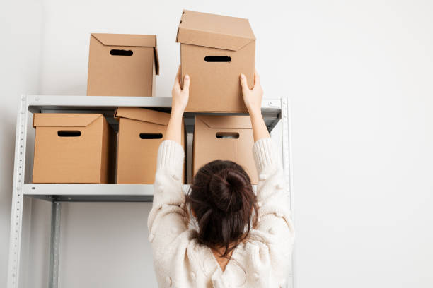 Woman put a cardboard box on a shelf of a rack in warehouse. Clean up and organize a pantry concept.  rear view. Woman put a cardboard box on a shelf of a rack in warehouse. Clean up and organize a pantry concept.  rear view. storage room stock pictures, royalty-free photos & images