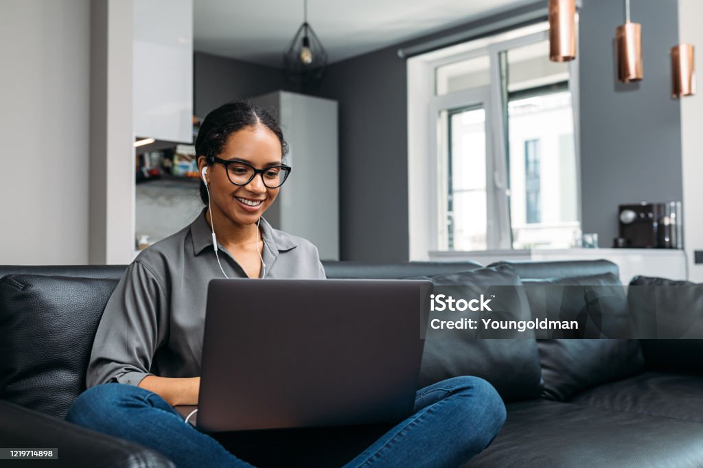 Young woman video calling using a laptop sitting on a sofa wearing earphones Working At Home Stock Photo