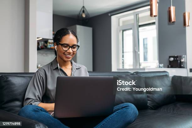Young Woman Video Calling Using A Laptop Sitting On A Sofa Wearing Earphones Stock Photo - Download Image Now