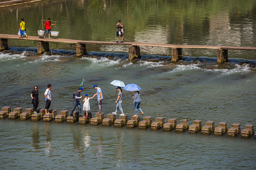 Feng Huang, China -  August 2019 : People crossing water on stepping stones on Tuo Tuojiang river, flowing through the centre of Fenghuang Old Town, Hunan Province