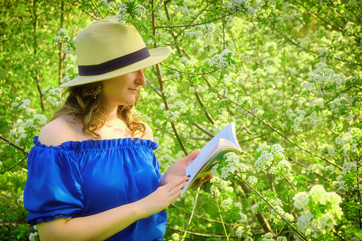 Young woman in a straw hat reading a book against the background of flowering trees in spring