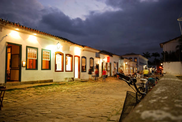 Cobblestone street in Paraty, Brazil, captured at night with warm lighting reflecting off the wet stones. The low-angle perspective emphasizes the historic texture of the street and colonial buildings. A bicycle rests against a whitewashed wall, and puddles on the street create mirror-like reflections of the glowing lamps and façades.