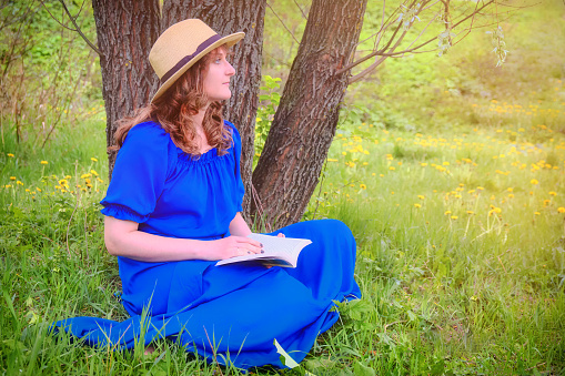 A woman in a blue dress reading a book sitting near a tree. Girl in a straw hat in the summer forest