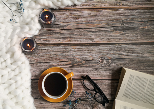 White knitted blanket , coffee cup , candles , reading glasses and open book on wooden surface. Top view , from above , flat lay. Still life, coziness , relaxing at home concept.