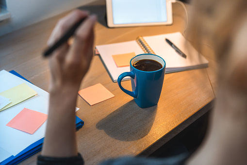 Woman working in her home office during pandemic quarantine. Close-up of a desk with coffee mug, digital tablet, notebook, folder, and papers.