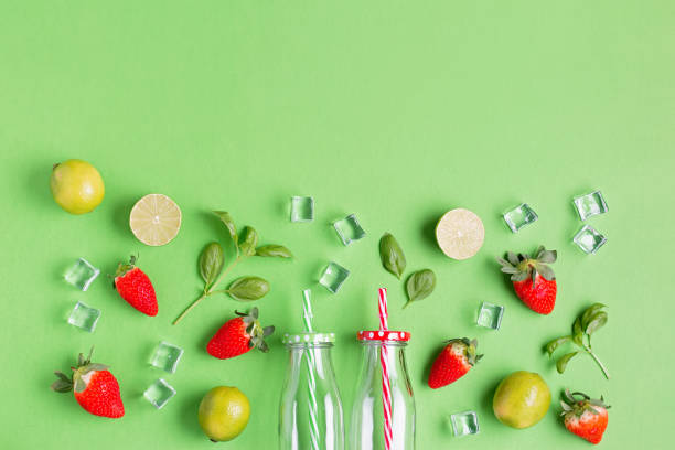 flat lay com ingredientes para a bebida refrescante de verão ou smoothie, cubos de gelo, ervas e potes de vidro no fundo verde, vista superior. morango, limão, gelo e manjericão para delicioso coquetel de verão - strawberry mocktail smoothie cocktail - fotografias e filmes do acervo