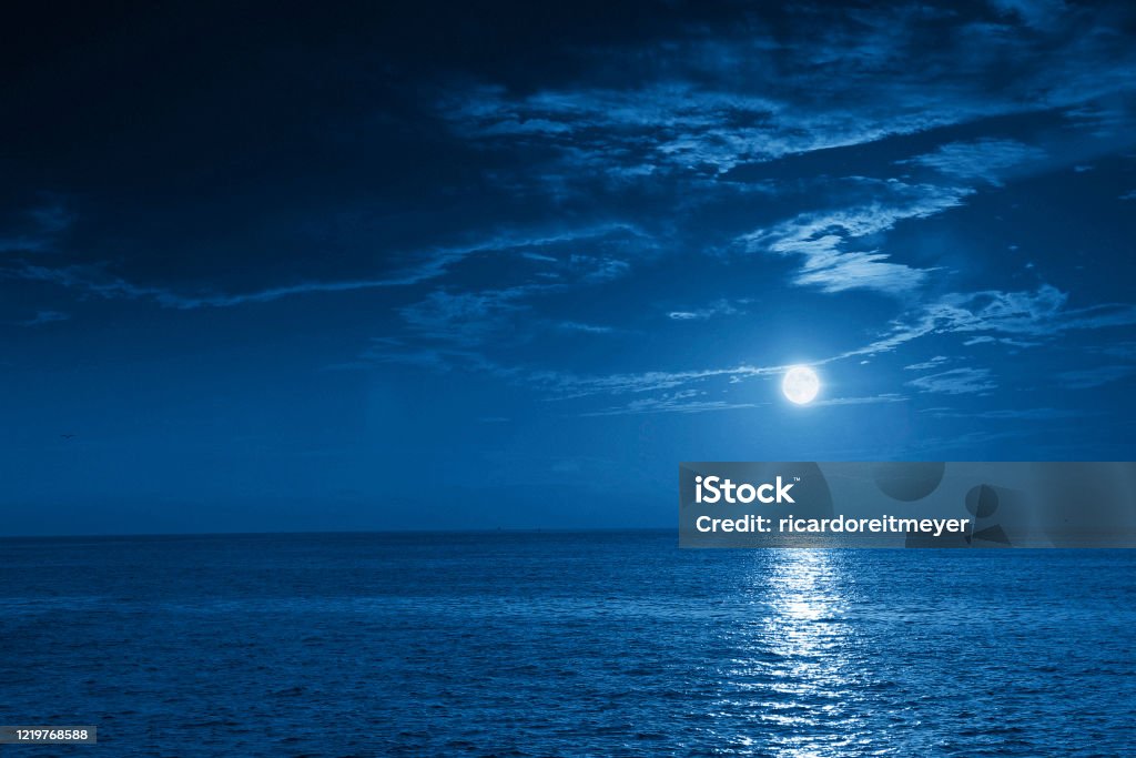 Bright Full Blue Moon Rises Over A Calm Ocean View This photo illustration of a deep blue moonlit ocean at night with calm waves would make a great travel background for any coastal region or vacation, emphasizing the beauty of the night time ocean or sea. Night Stock Photo