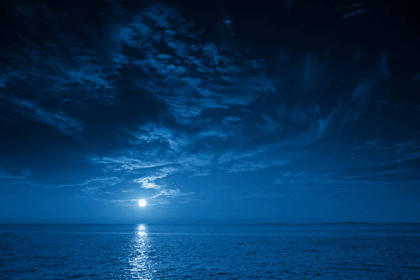 Bright Full Blue Moon Rises Over A Calm Ocean View This photo illustration of a deep blue moonlit ocean at night with calm waves would make a great travel background for any coastal region or vacation, emphasizing the beauty of the night time ocean or sea. atlantic ocean photos stock pictures, royalty-free photos & images