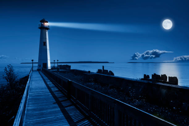 This is a beautiful photo illustration of a dramatic night time scene with a large blue moonrise in a clear sky on a ocean pier with a brightly lit lighthouse beacon and calm ocean waters.