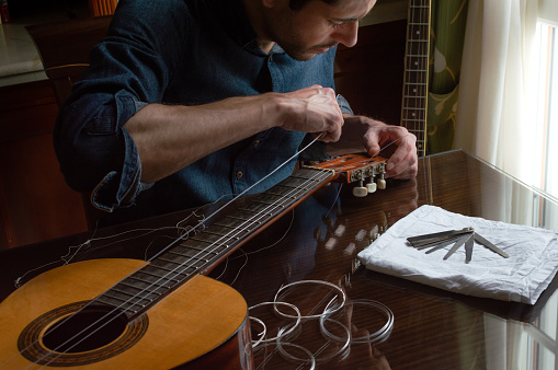 Focus man changing the strings of a guitar at home. Wooden table with new strings in classic indoor place.
