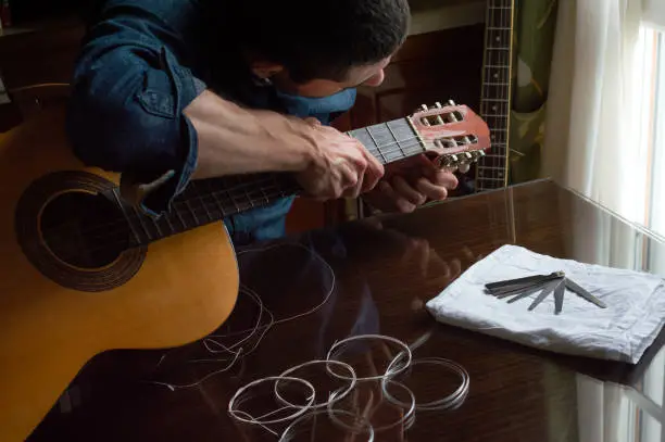 Photo of Man in shirt preparing and changing the strings of a spanish guitar at home near to a window. Wooden table with new strings in classic indoor place.