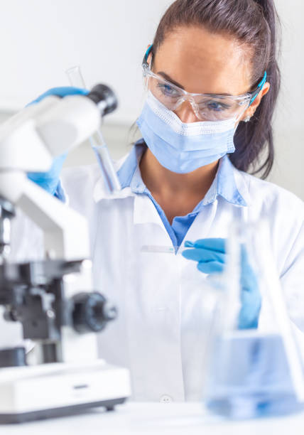 mujer con capa blanca, gafas protectoras y máscara facial mira el tubo de ensayo en una mano, sosteniendo un portaobjetos de microscopio en la otra mano. coronavirus covid-19 concepto. - science innovation microscope healthcare and medicine fotografías e imágenes de stock