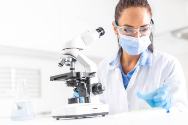istock Female lab technician in protective glasses, gloves and face mask sits next to a microscope and conical flask, looking aside on the desk. 1219770491