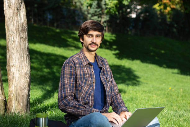 istock Young bearded brunet student with mustache sitting with a laptop in a park on grass 1219777073