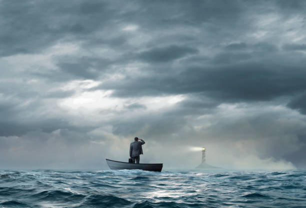 Businessman Looks At Lighthouse While Stranded On Boat A beacon from a lighthouse beckons a stranded businessman as he stands in a small boat that floats under an ominous sky and choppy waters. lost stock pictures, royalty-free photos & images