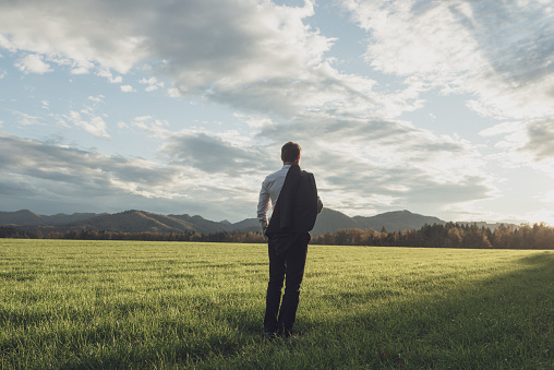 View from behind of a young businessman with his suit jacket over his shoulder, standing in beautiful green meadow under cloudy evening sky.