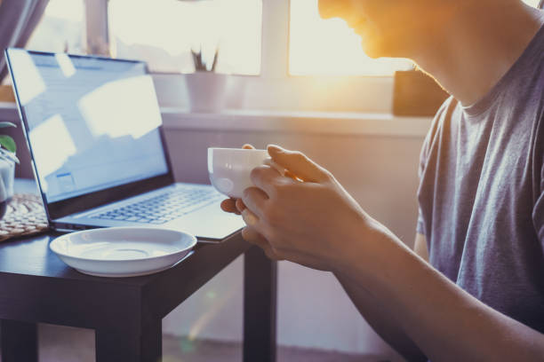 Young man working on laptop sitting near coffee table with cup of hot drink tea. Workplace near window on sutset or early morning. Working at home. Back light, soft selective focus. Copy space joven que trabaja en la computadora portátil sentado cerca de la mesa de café con una taza de té de bebida caliente. lugar de trabajo cerca de la ventana en sutset o temprano en la mañana. trabajando en casa. luz trasera, enfoque selectivo suave. copia