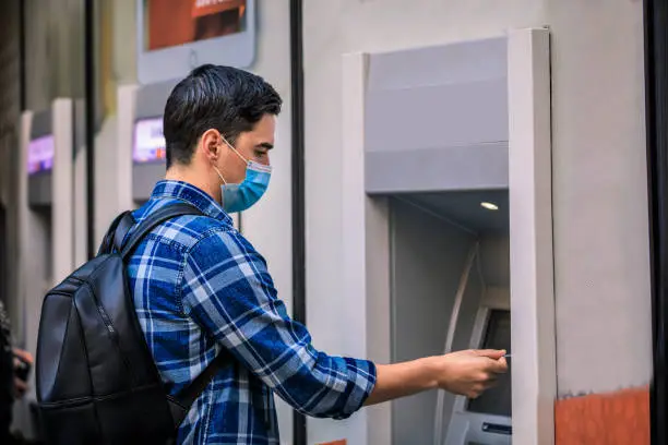 Man using a credit card in an atm for cash withdrawal. Concept, diseases, viruses, allergies, air pollution. Portrait of young man wearing a protective mask, walking in the city.The image face of a young man wearing a mask to prevent germs, toxic fumes, and dust. Prevention of bacterial infection Corona virus or Covid 19 in the air around the streets and gardens.