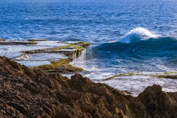 Tidepool on overhanging ledge, water cascading back into the sea, Devil's Tear, Nusa Lembongan, Bali, Indonesia. Wave with white crest, blue ocean beyond.