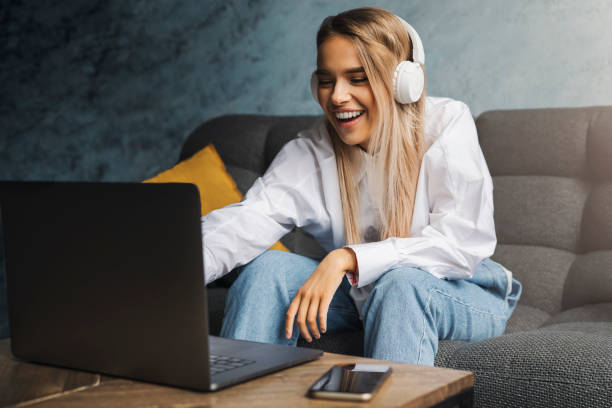 cheerful girl in headphones sitting on sofa in front of laptop. entertainment online content for streaming at affordable - financial figures audio imagens e fotografias de stock
