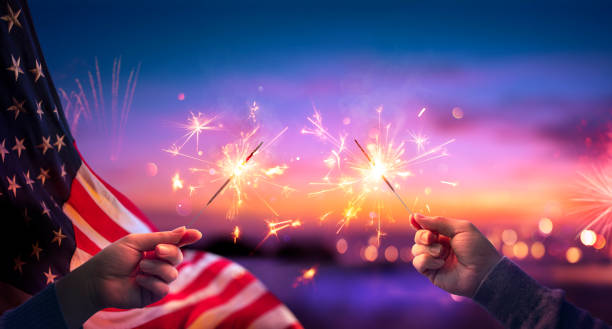 usa feier mit händen halten sparklers und amerikanische flagge bei sonnenuntergang mit feuerwerk - 4th of july stock-fotos und bilder