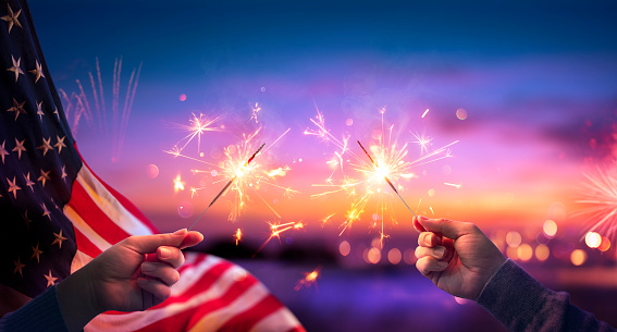 Celebración de Usa con las manos sosteniendo destellos y bandera americana al atardecer con fuegos artificiales photo