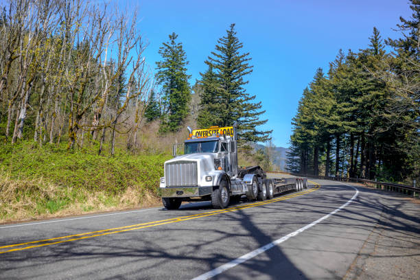 istock Powerful big rig white semi truck with oversize load sign on the roof transporting empty step down semi trailer for carry heavy-duty cargo running on the road with forest on the hill 1219817976