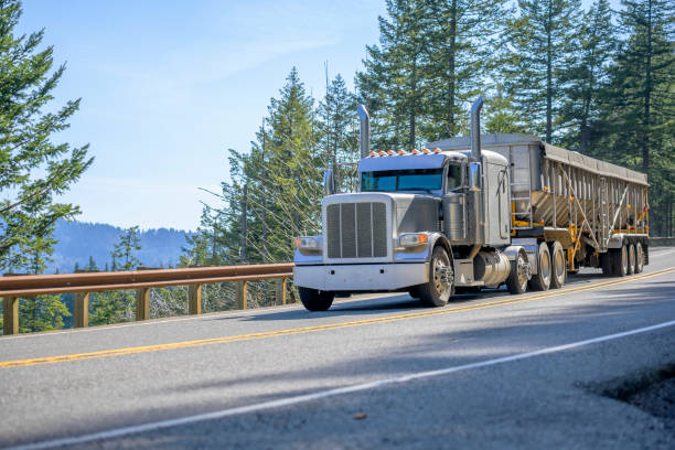 istock Gray classic big rig semi truck with vertical pipes transporting cargo in covered bulk semi trailer running on the mountain road with trees on the side 1219818012