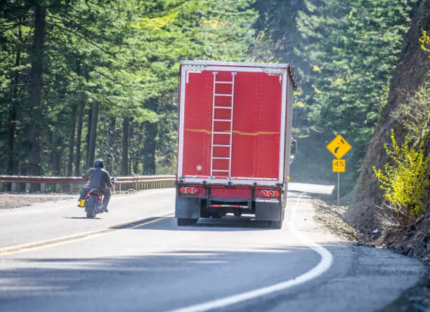 Photo of Motorcyclist overtaking a big rig semi truck with red bulk semi trailer on the winding mountain road in prohibited no-overtaking zone without a secure road view