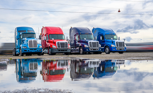 Powerful clean big rigs semi trucks tractors with high cab and rest compartment for truck drivers resting standing on the warehouse parking lot waiting for loaded semi trailer for next delivery