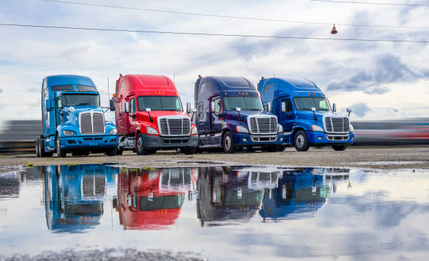 DIfferent big rigs semi trucks standing in row on the parking lot with reflection in water after the rain Powerful clean big rigs semi trucks tractors with high cab and rest compartment for truck drivers resting standing on the warehouse parking lot waiting for loaded semi trailer for next delivery commercial land vehicle stock pictures, royalty-free photos & images