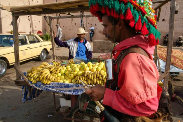 Marrakesh, Morocco - February 5, 2016: A local man sells fruit outside the walls of Marrakesh’s Medina by Bab Doukkala, as a Guerrab (Water Carrier) walks by in traditional red clothing.