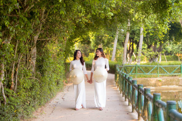 Two Vietnamese girls wearing a white Ao Dai and holding a conical hat at Hoang Tru, Kim Lien, Nam Dan. The Ao dai ( long-dress Vietnamese) is traditional costume of Vietnamese woman Two Vietnamese girls wearing a white Ao Dai and holding a conical hat at Hoang Tru, Kim Lien, Nam Dan. The Ao dai ( long-dress Vietnamese) is traditional costume of Vietnamese woman ao dai stock pictures, royalty-free photos & images