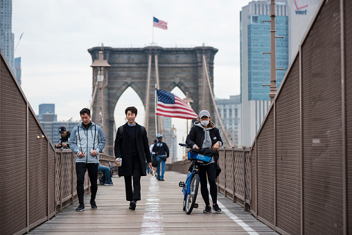 New York City, USA - March 20, 2020: Three people, one wearing a surgical mask, walk across the Brooklyn Bridge in the direction of Brooklyn. In the background, a man holding an American flag walks in the direction of Manhattan.