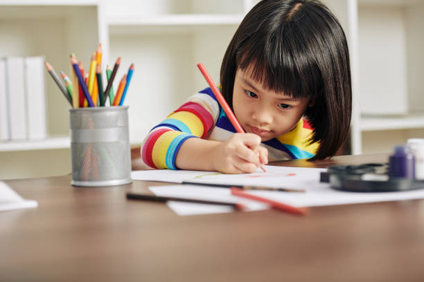 Drawing little girl Adorable talented Asian girl sitting at desk at home and drawing with red pencil coloring photos stock pictures, royalty-free photos & images